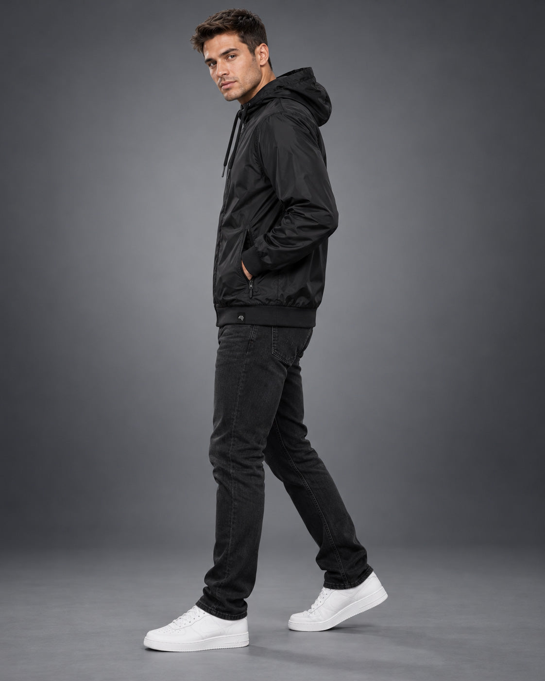 Herren-Windjacke am Model im Fotostudio, schwarzer Nylonstoff mit roten Reißverschlüssen und Kapuzenkordeln, breiter schwarzer Rippbund und sportliche Passform